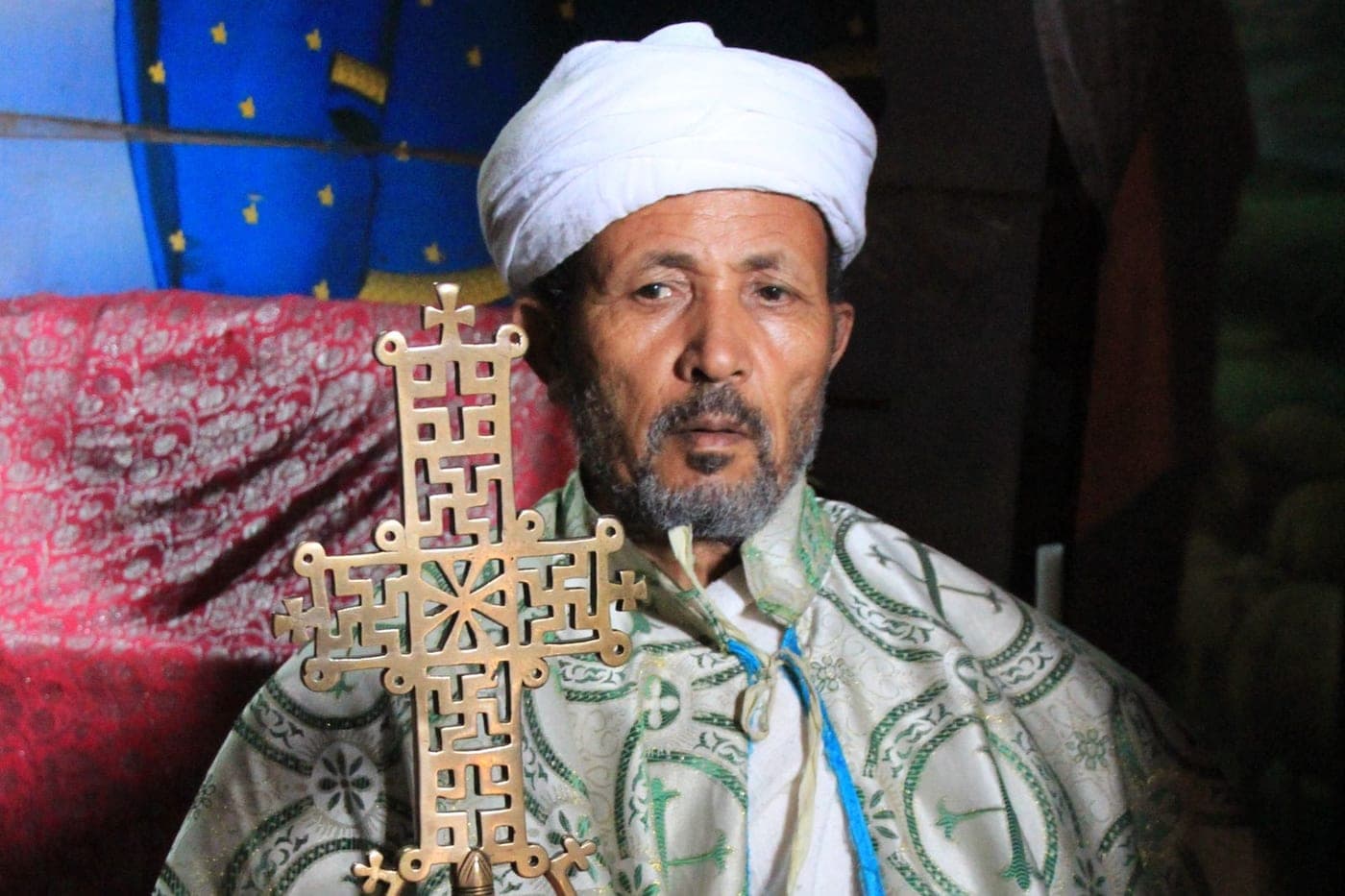 An Ethiopian Orthodox priest at Lalibela in embroidered vestments and a white turban, holding a large brass processional cross.