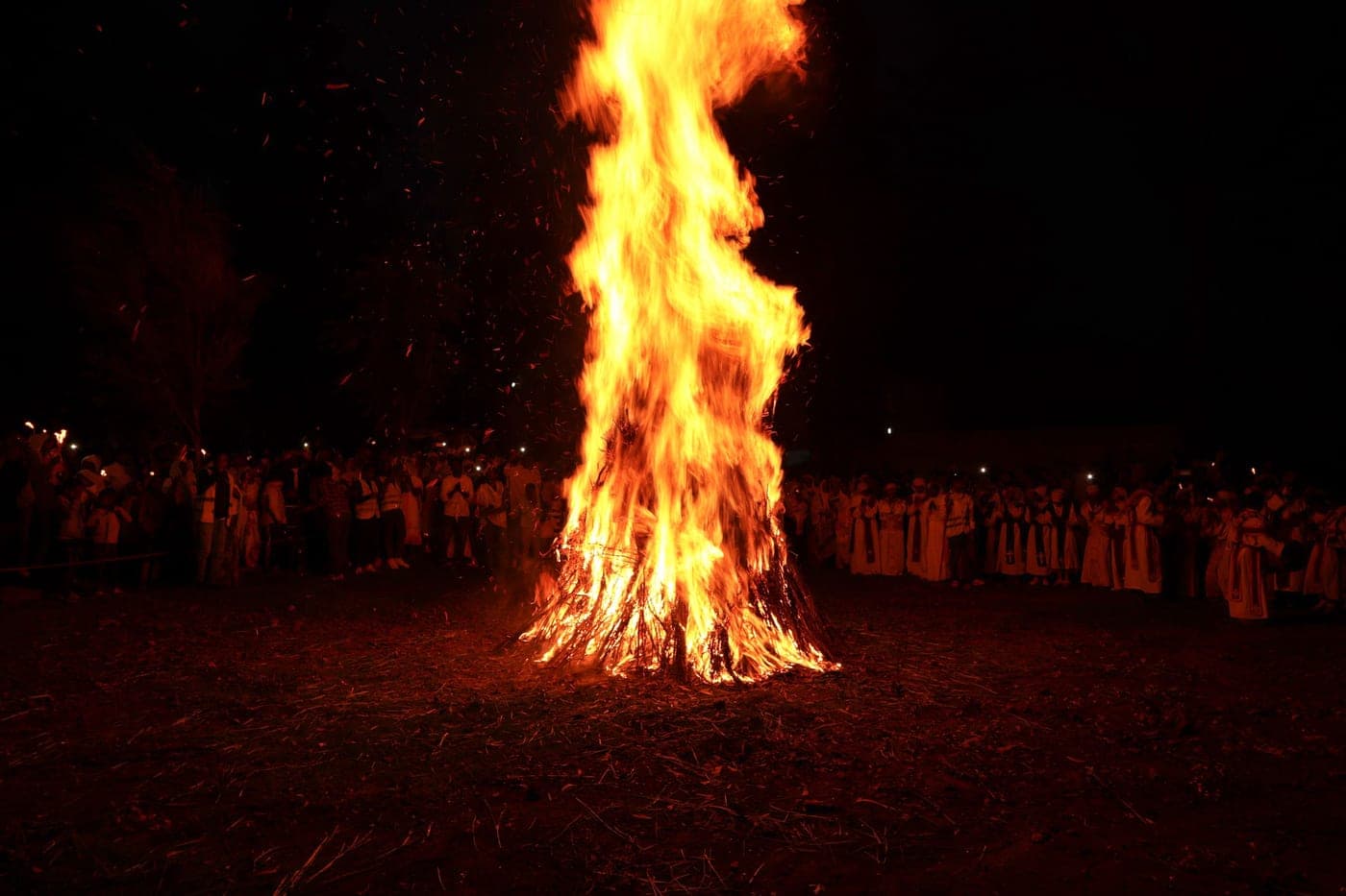 The Demera bonfire burning at night, ringed by a crowd holding candles, with clergy in ceremonial robes standing at the right.