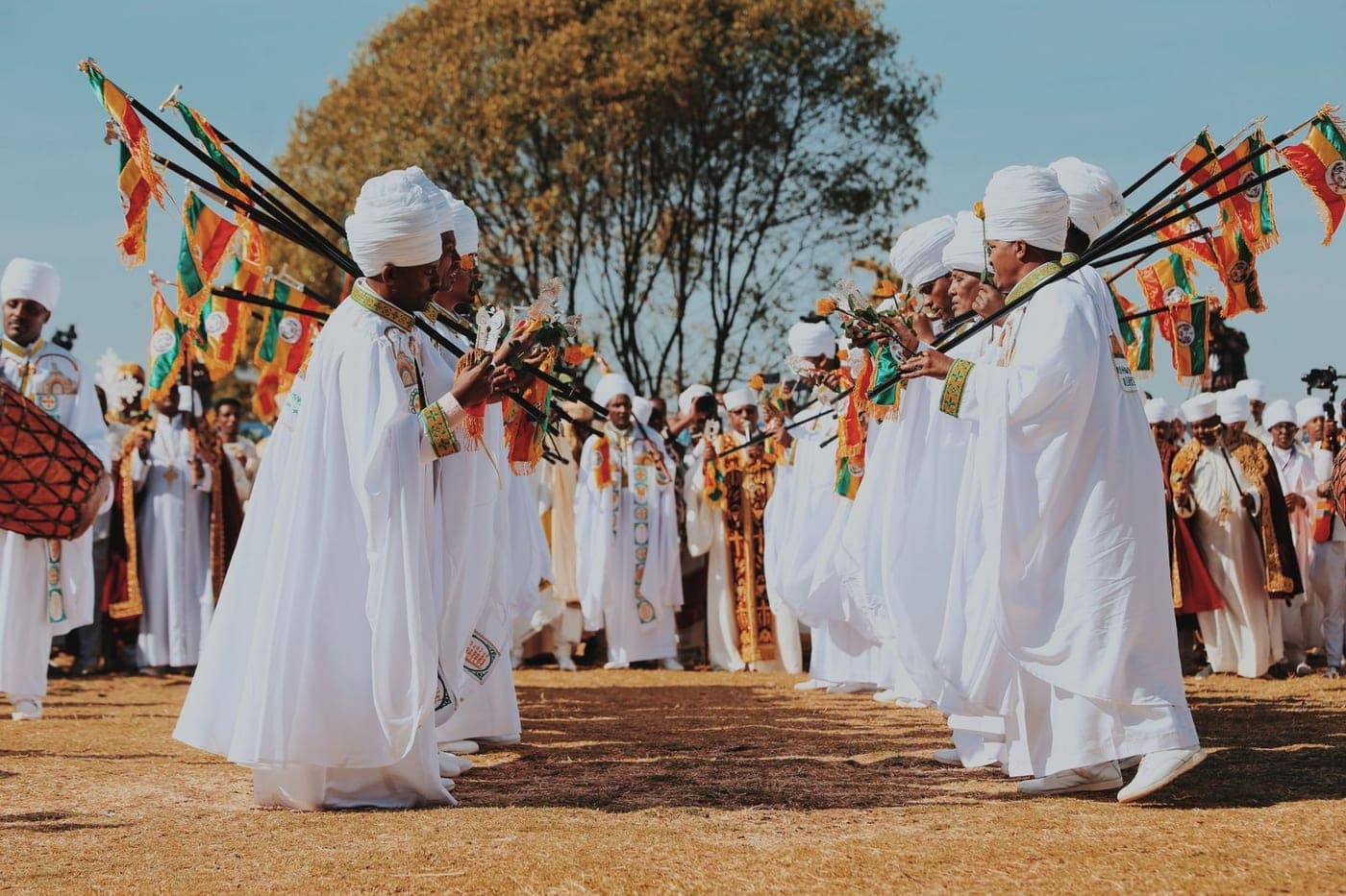 Ethiopian Orthodox clergy in white robes facing each other with church flags and a drum, on dry highland ground under a clear sky.