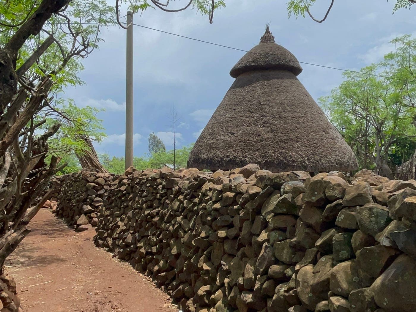 A red earth lane running beside a dry-stone wall in a Konso settlement, with a tall conical thatched communal house rising behind it.