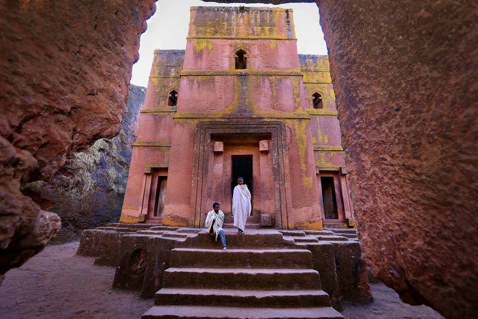 Two men in white shawls on the steps of a rock-hewn church at Lalibela, its facade cut down into the surrounding stone and framed by the walls of the trench.